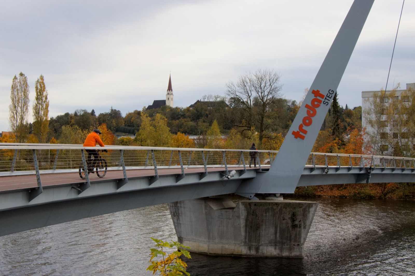 Radfahrer quert den Trodatsteg über die Traun bei Wels, im Hintergrund die Kirche von Thalheim.